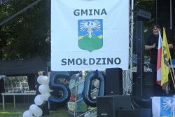 Biały baner z napisaem "Gmina Smołdzino" i logotypem gminy, którym jest bielik na tle zielonego wzgórza, z żółtym paskiem i niebieskim tłem.